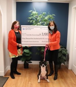 Two women stand on either side of a large check representing the donation eFS is making to the Great Lakes Humane Society. Patty Cake, the dog, is sitting in between them. Both humans are wearing face masks.