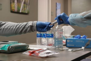 Photo shows two hands in gloves handing off a tube to one another where the nasal swab will be placed for testing.