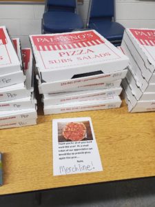 A stack of pizza boxes is shown on a table with a paper sign in front of the boxes that reads, "Thank you for all of your hard work this year! As a small token of our appreciation we would like to provide pizza again this year. Nate, Merchline!"