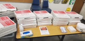 A long rectangle table is shown with several stacks of pizza boxes. Also on the table is hand sanitizer, gloves, plates, napkins and a note of thanks from Merchline.