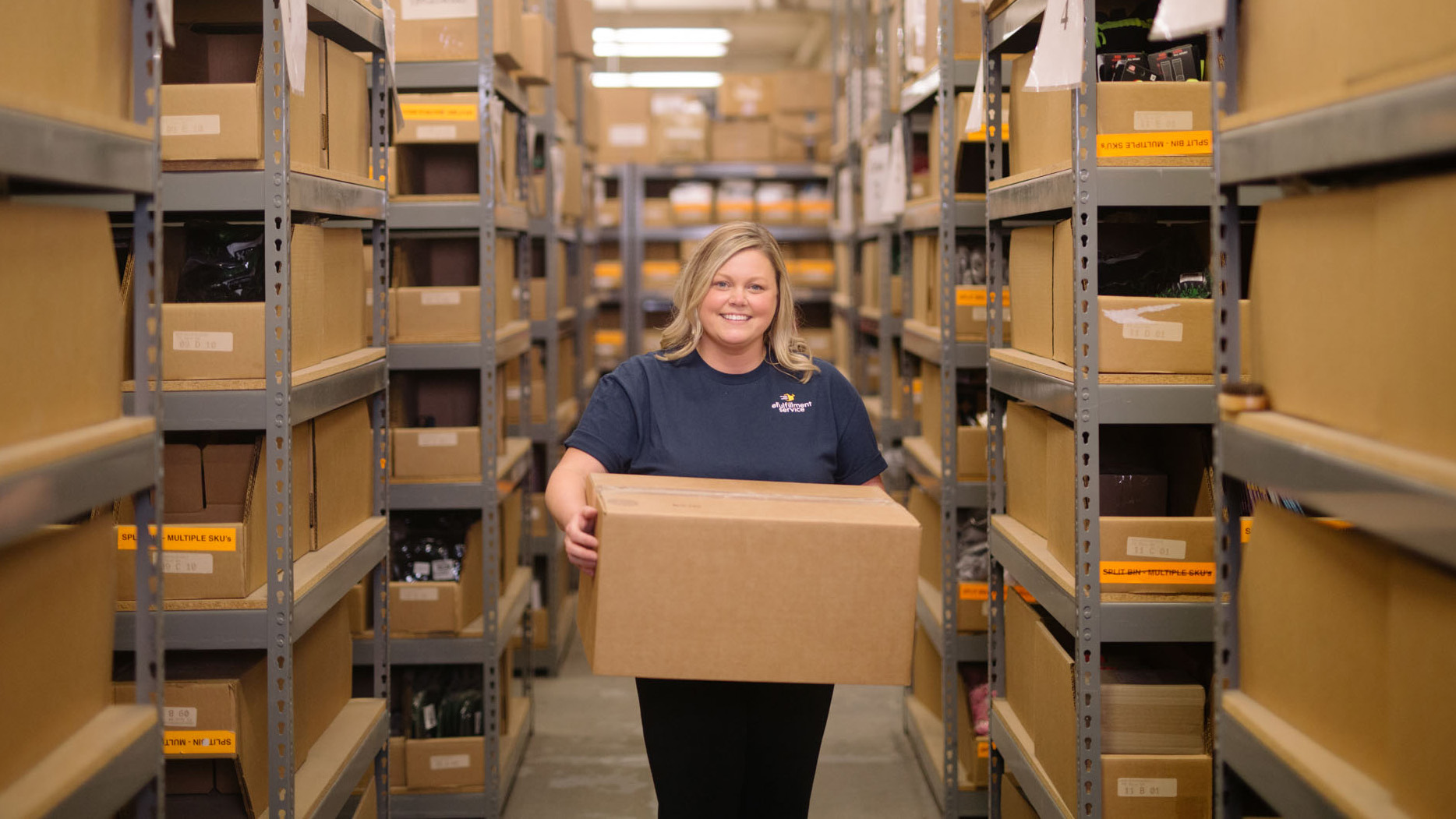 picture of a women warehouse worker holding a box among many shelves in a warehouse