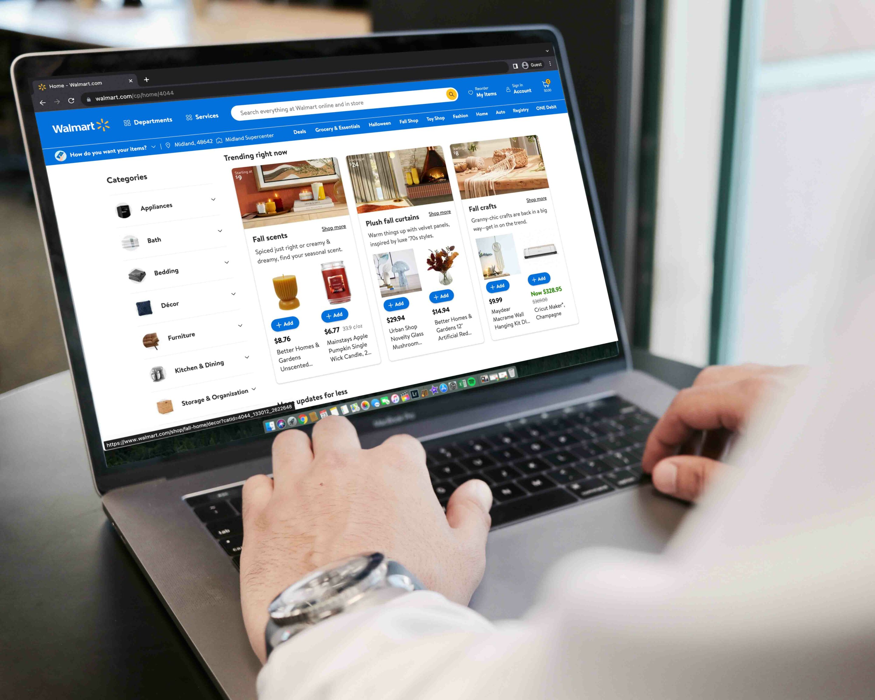 A person browsing the Walmart website on a laptop. The website displays various product categories like Appliances, Bath, Bedding, and Decor, along with featured products such as fall scents and home decor items.