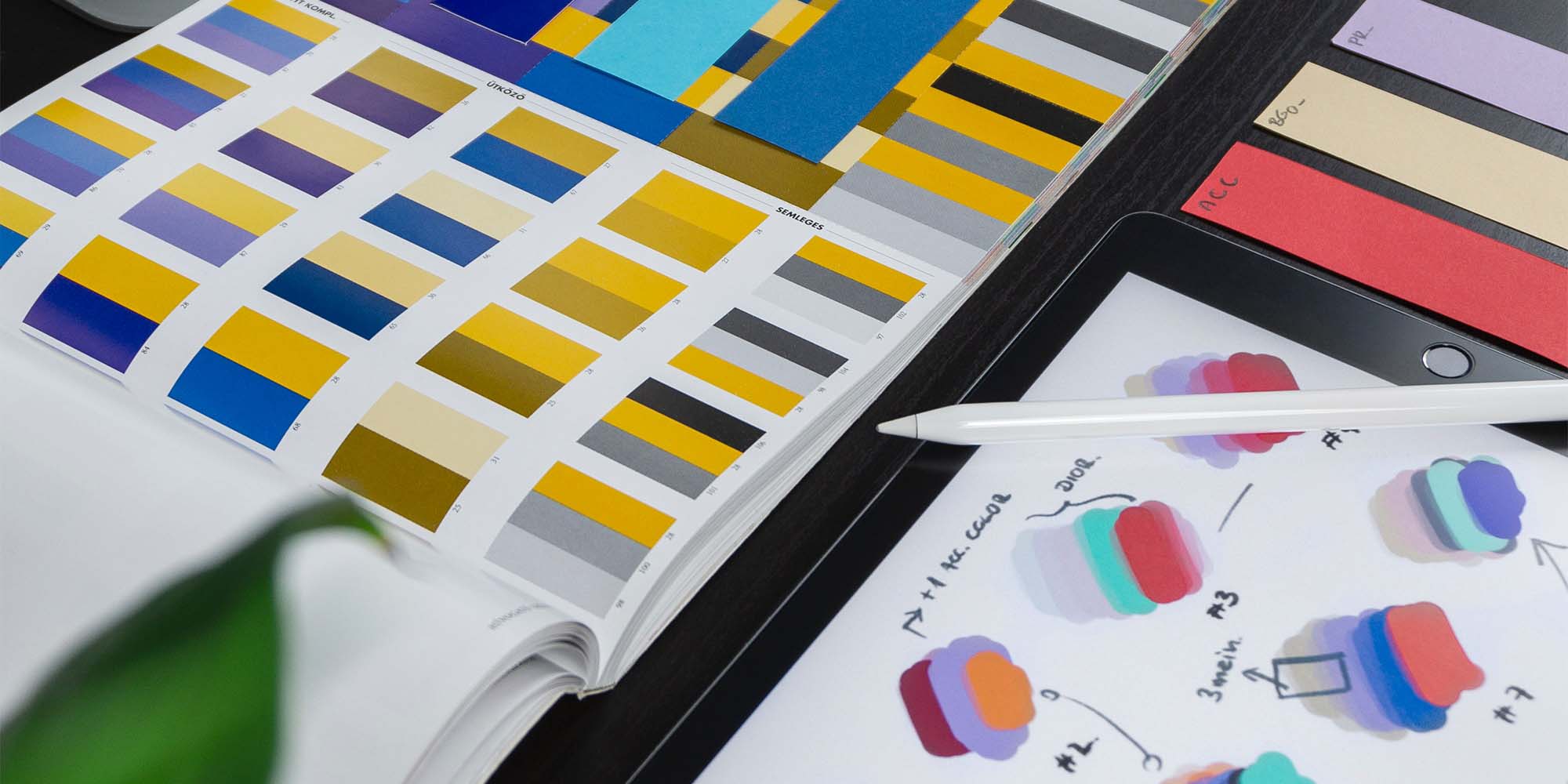 Designer's workspace with an open color swatch book, an iPad displaying graphic designs, and samples of colored paper on a dark table.