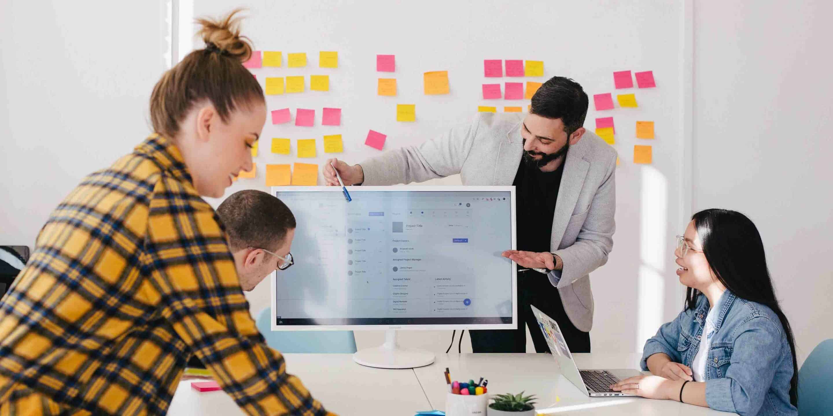 Team collaboration in a modern office with a man presenting on a screen to colleagues, colorful sticky notes in the background